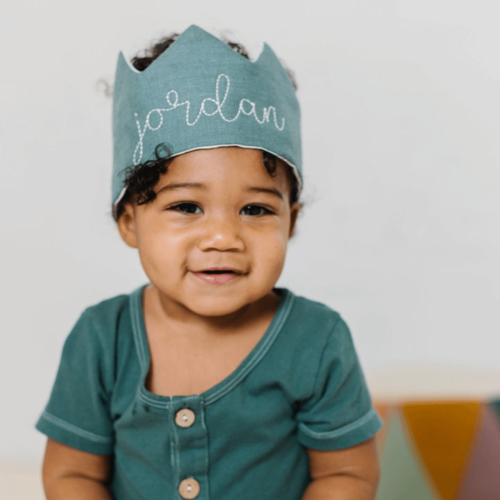 Baby with green crown that reads 'Jordan' sitting on yellow chair