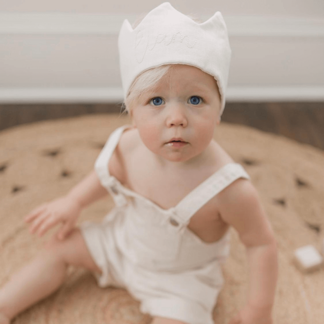 Blonde baby sitting on floor wearing a personalized white linen birthday crown