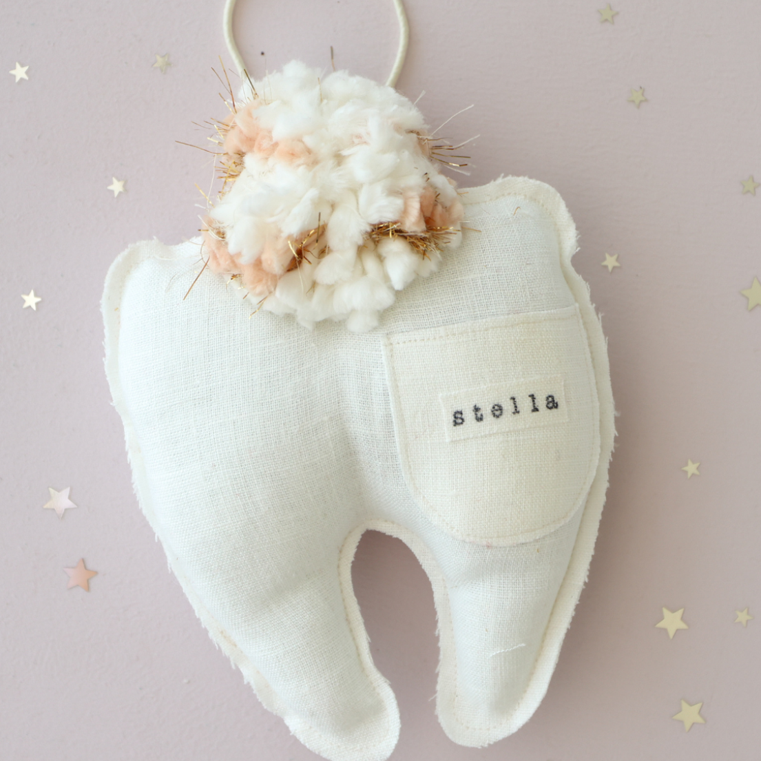 Close-up of white linen tooth-shaped pillow with coral pom pom and personalized name 'Stella' on front pocket