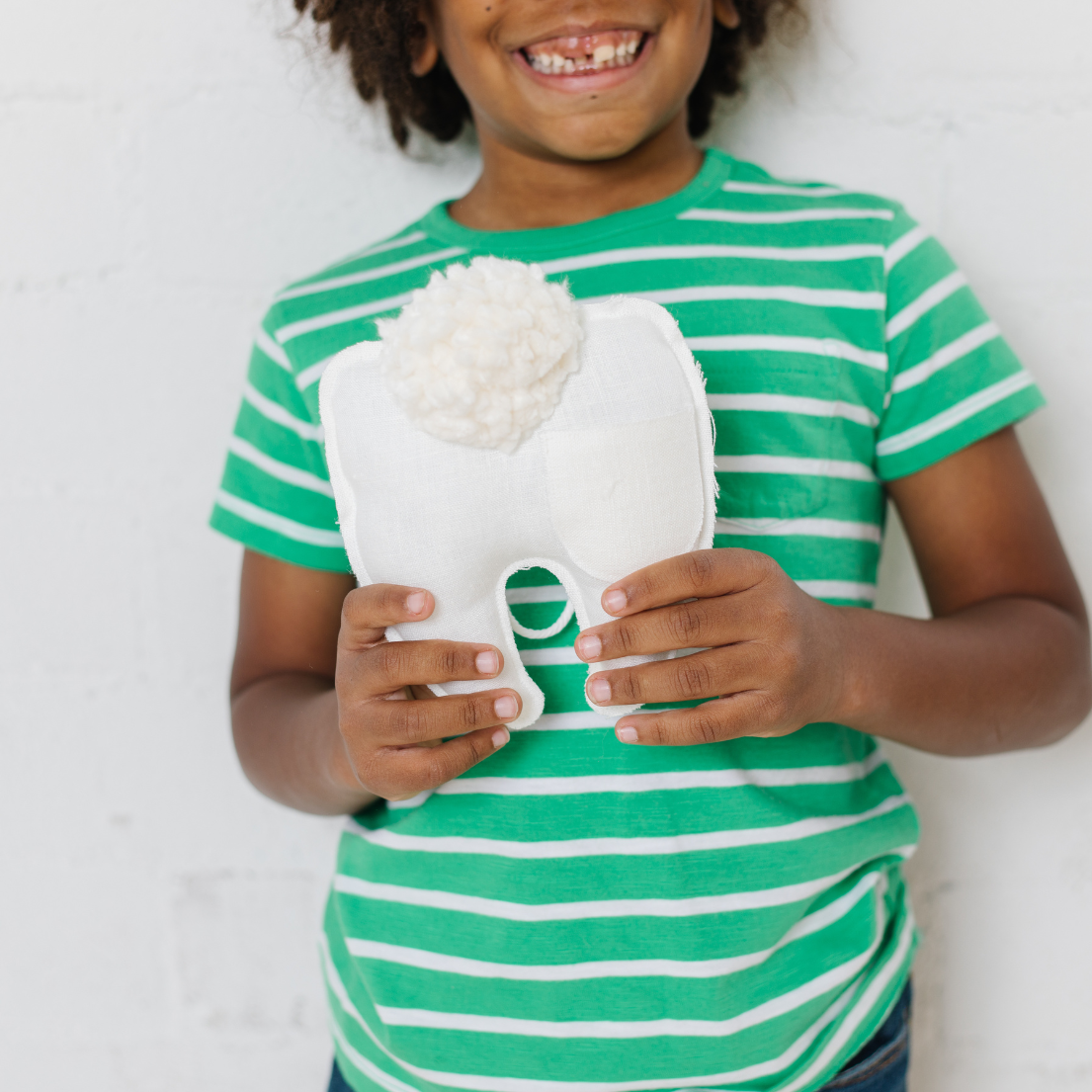 Smiling child in green striped shirt holding tooth-shaped linen pillow with pom pom