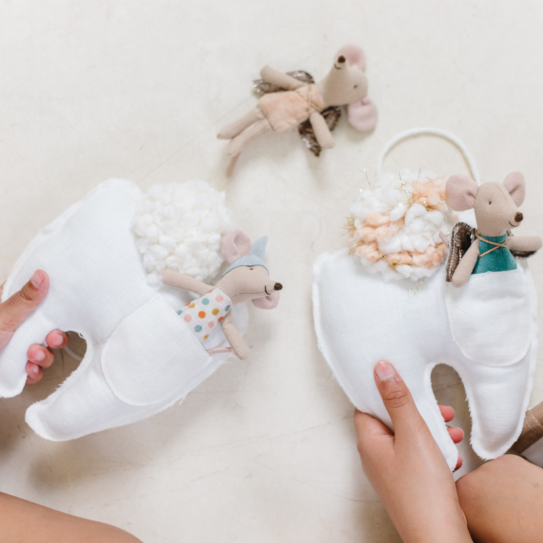 Flat lay of two white linen tooth pillows with coral and neutral pom poms, child's hand reaching