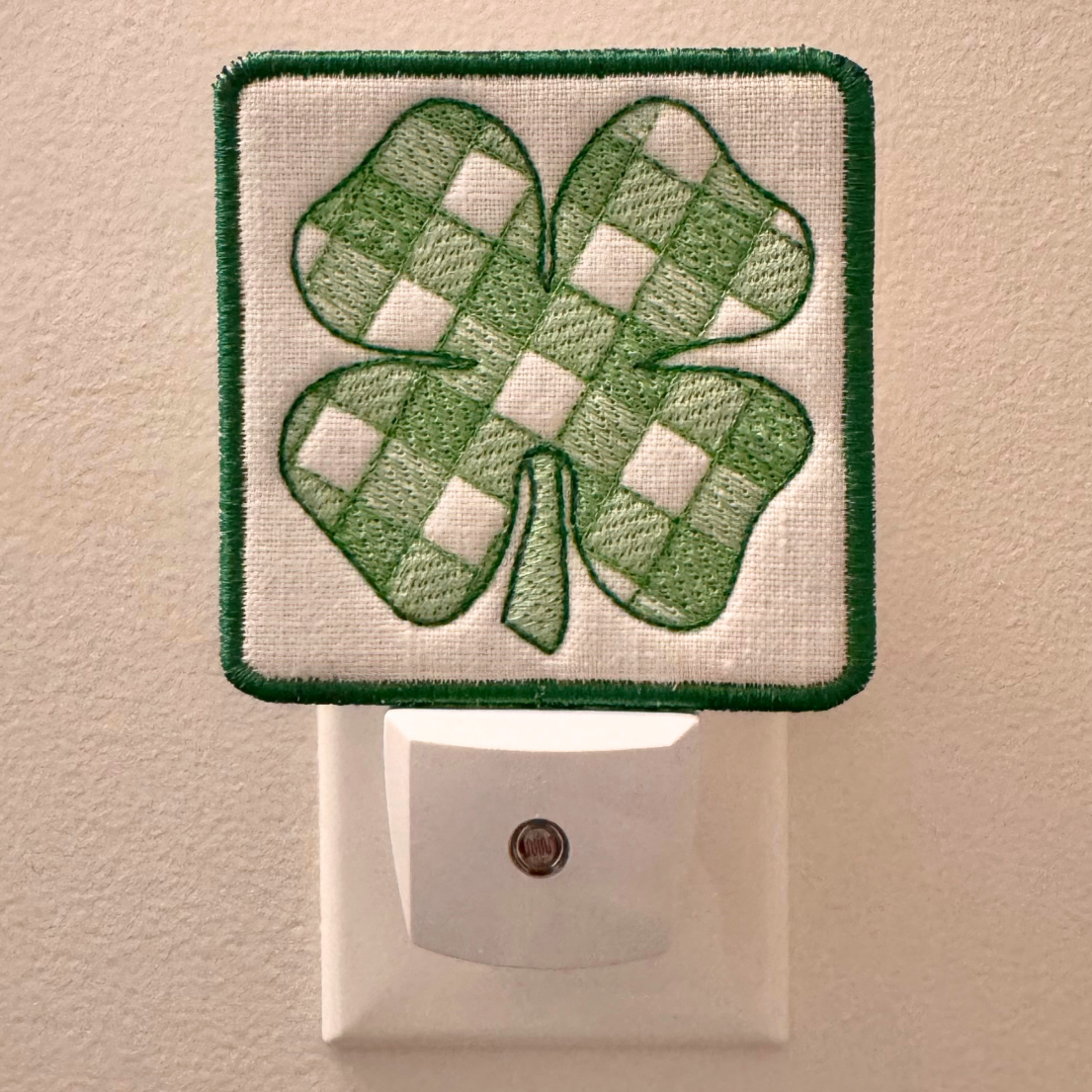 A square white night light patch with a green embroidered shamrock on the front, shown plugged into a white wall outlet against a beige wall.