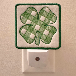 A square white night light patch with a green embroidered shamrock on the front, shown plugged into a white wall outlet against a beige wall.