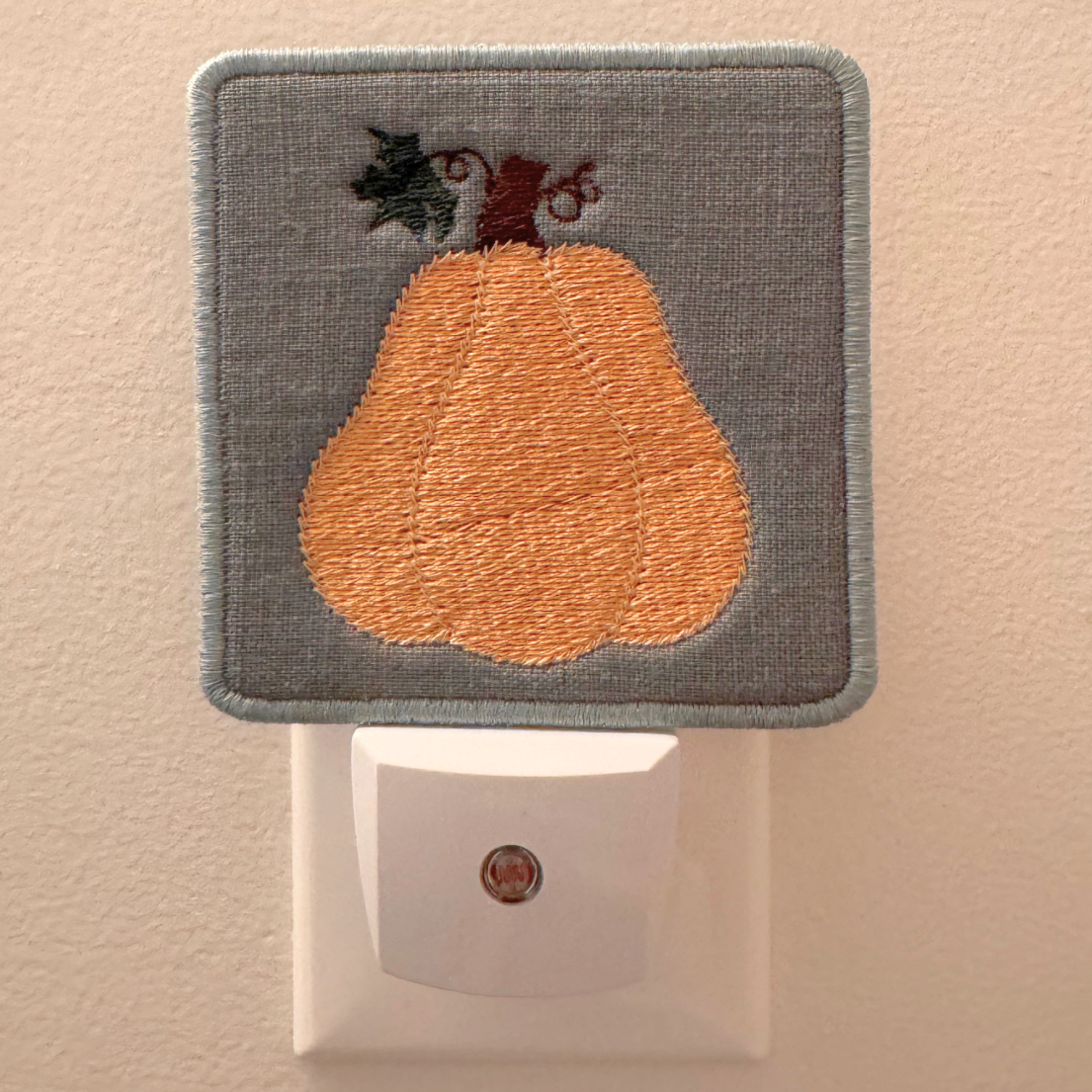 An embroidered night light cover featuring a peach-colored pear on a gray fabric background, shown plugged into a white wall outlet against a beige wall.