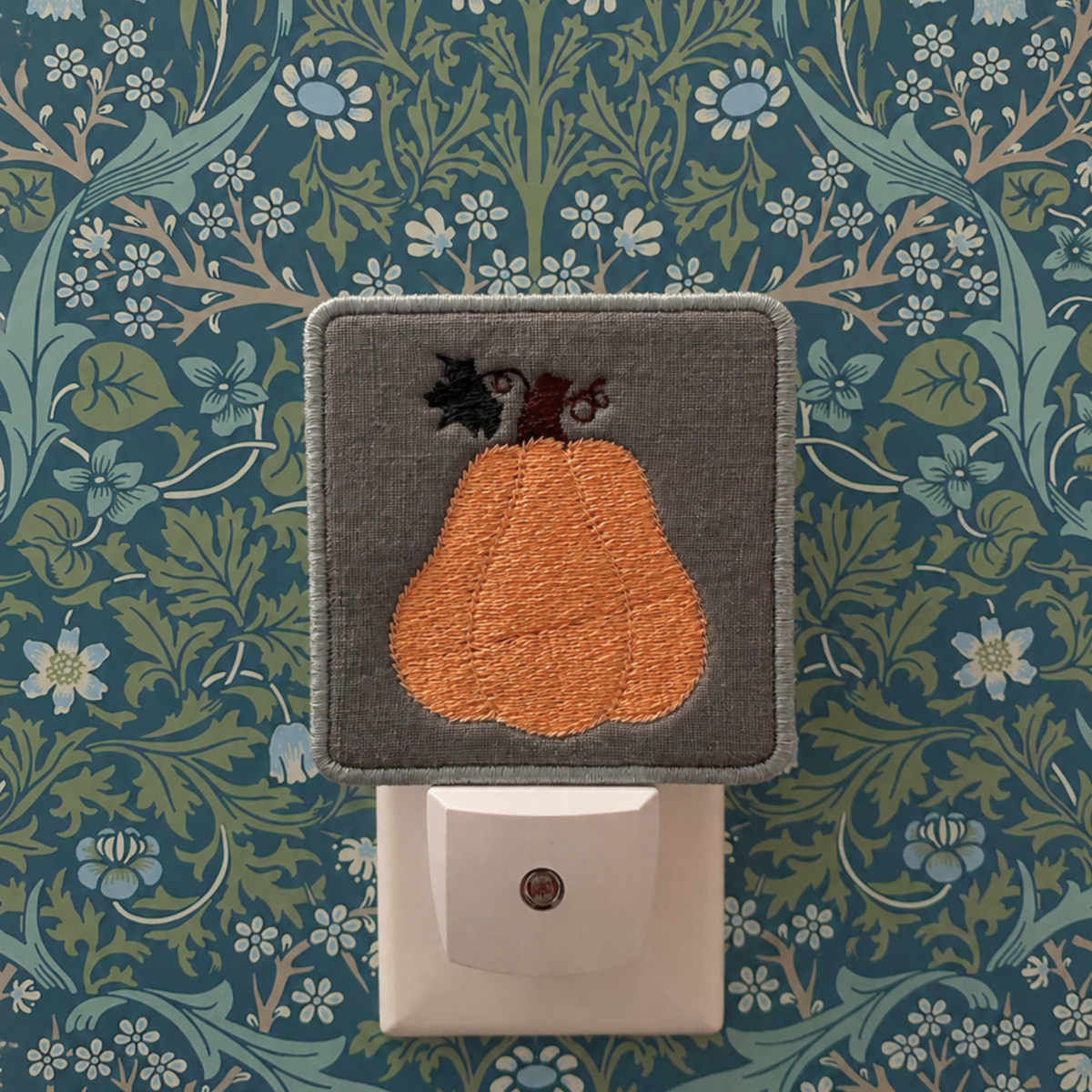 A square gray fabric night light cover featuring an embroidered orange pear with a dark stem, shown plugged into a white wall outlet against a blue and green floral wallpaper background.