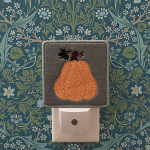 A square gray fabric night light cover featuring an embroidered orange pear with a dark stem, shown plugged into a white wall outlet against a blue and green floral wallpaper background.