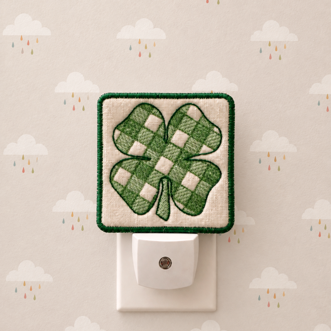 A square quilted St. Patrick's Day night light with a green and white checkered shamrock on a cream fabric background, shown plugged into a white outlet against a patterned wallpaper with clouds and raindrops.