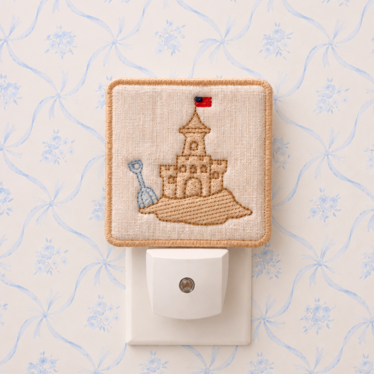 A square fabric night light cover with a stitched sandcastle design and a small flag, shown plugged into a white wall outlet against a light patterned wallpaper background.