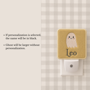 A personalized ghost night light shown mounted on a white plug. The fabric cover is mustard yellow with an embroidered white ghost and the name 'Leo' in black.
