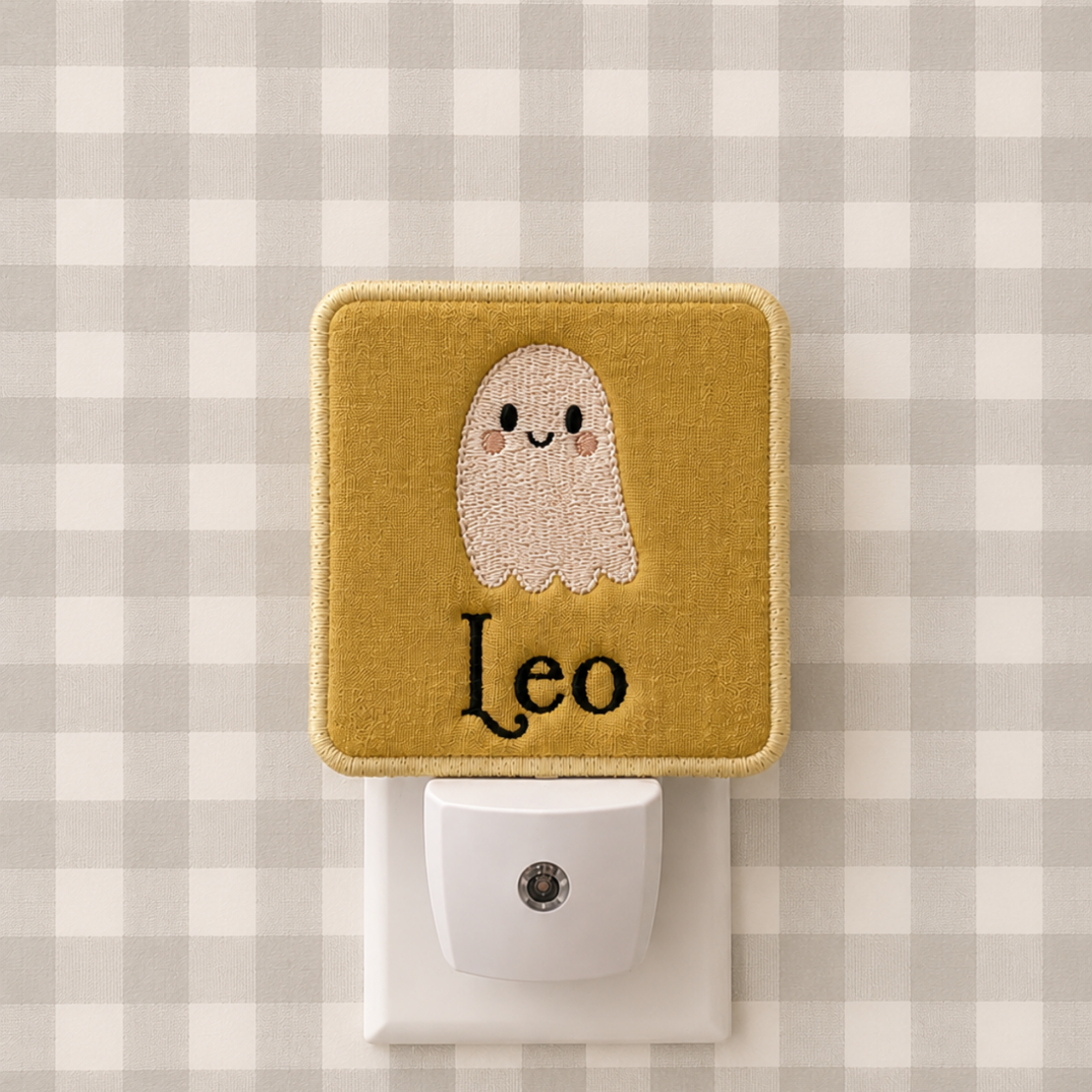 A square fabric night light plug-in with a mustard yellow border and a cute embroidered ghost face above the name 'Leo' in black script. The white plug is inserted into a white wall outlet against a gray and white gingham patterned background.