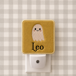 A square fabric night light plug-in with a mustard yellow border and a cute embroidered ghost face above the name 'Leo' in black script. The white plug is inserted into a white wall outlet against a gray and white gingham patterned background.