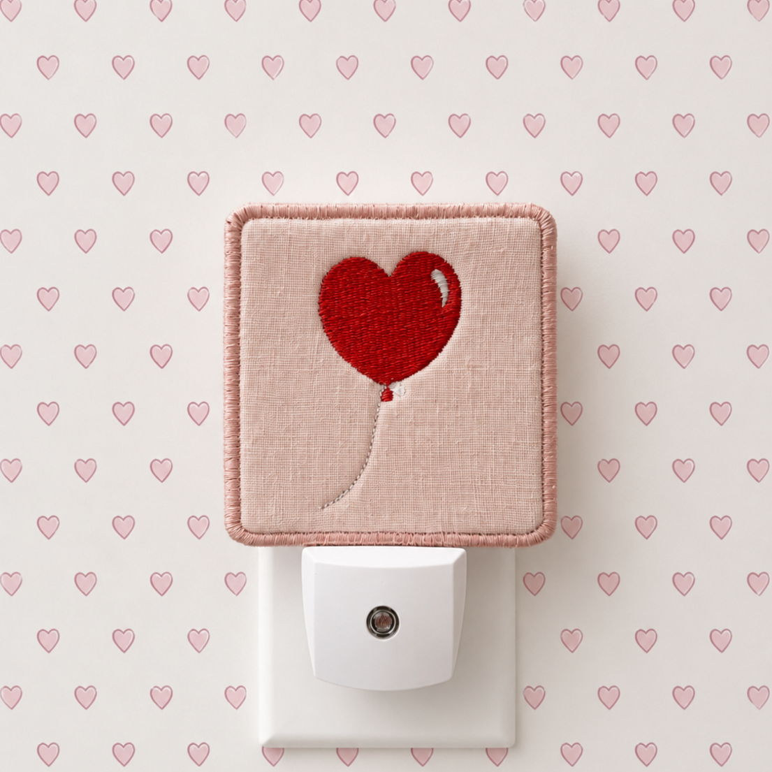 A square pink fabric night light cover with a pink heart balloon design, shown plugged into a white outlet against a patterned wallpaper background.