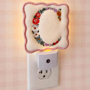 A decorative embroidered letter C night light plug-in is shown plugged into a wall outlet. The letter C is embroidered in a floral wreath design with red, orange, green, and purple flowers on a cream fabric background, surrounded by a pink stitched border.