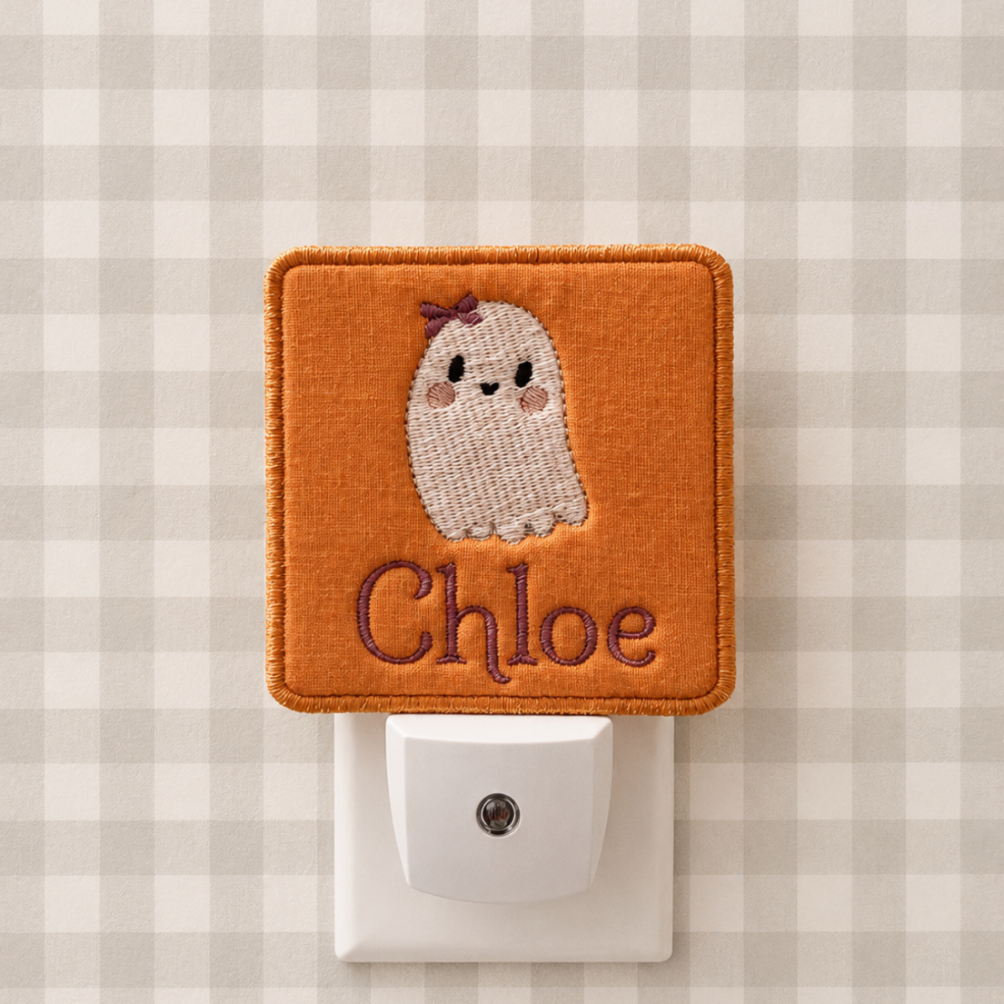 An orange embroidered night light cover on a white plug-in socket, featuring a cute white knitted ghost with a small purple bow and the name 'Chloe' in purple script. The background is a beige and white gingham pattern.