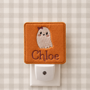 An orange embroidered night light cover on a white plug-in socket, featuring a cute white knitted ghost with a small purple bow and the name 'Chloe' in purple script. The background is a beige and white gingham pattern.