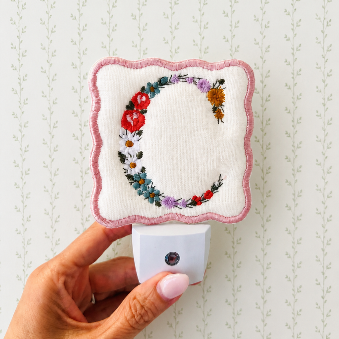 A hand holds a square white fabric night light cover with scalloped pink edges and an embroidered letter C in a floral wreath design, attached to a white night light plug against a pale patterned wallpaper background.