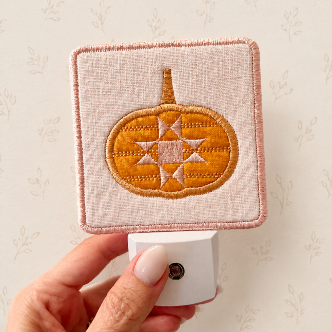 A hand holds a square fabric night light with a stitched orange pumpkin design on a pale pink background, against a light floral wallpaper backdrop.