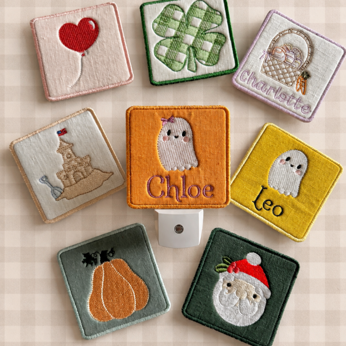 A set of personalized square fabric patchwork night lights arranged on a beige gingham background, with a small white plug-in holder in the center. The covers feature embroidered designs including a red heart, green shamrock, basket with the name Charlotte, sandcastle, a ghost with the name Chloe, a ghost with the name Leo, a pumpkin, and a Santa face on dark green, orange, yellow, and sage green linen.